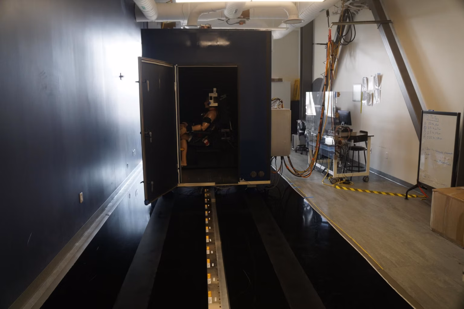 The Tilt Translation Sled laboratory: a dark enclosed cabin mounted on a long linear rail, doors open with an instrumented subject visible inside, control workstation and cabling to one side.